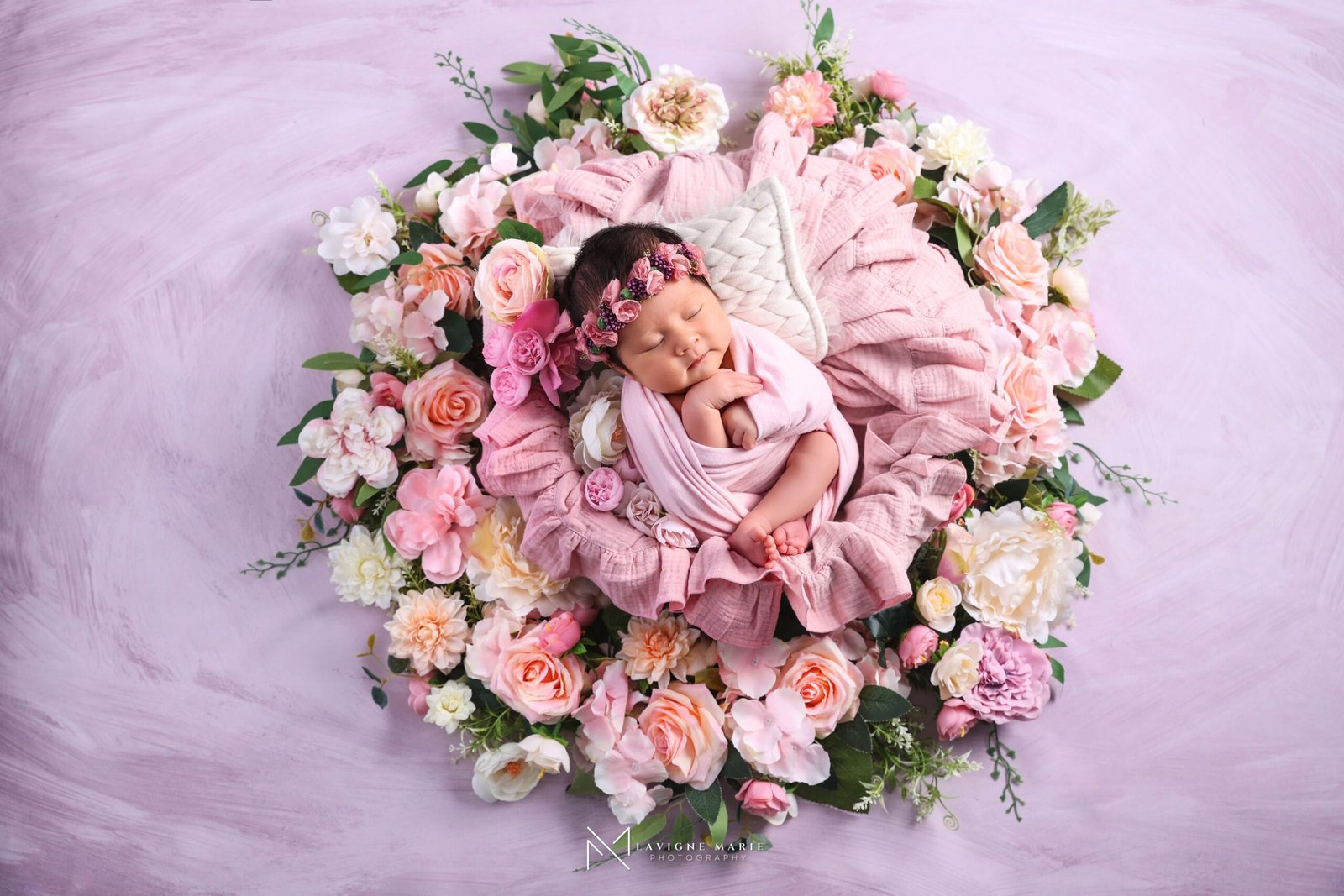 Sleeping newborn wrapped in pink muslin, wearing a floral headband with pink and purple flowers, surrounded by a white knitted angel wing prop, pink ruffled blanket, and a bouquet of pink, peach, and white flowers with green foliage against a light purple textured background