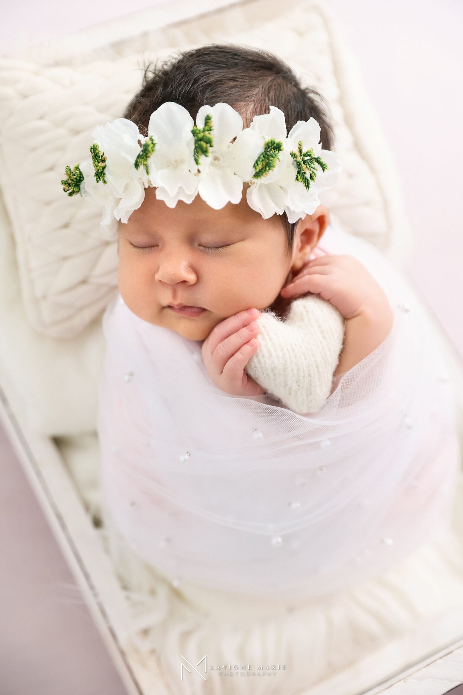 Sleeping newborn baby in a white wooden box, wearing a white flower headband with green centers and dark hair, eyes closed, holding a white knitted heart prop, wrapped in a sheer white fabric with pearl embellishments, surrounded by a white textured blanket