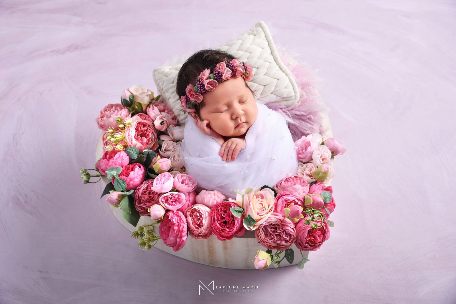 sleeping newborn wrapped in white sheer fabric, wearing a floral headband with pink flowers and purple berries, lying on a round wooden prop surrounded by pink and red roses, green foliage, a white textured pillow, pink fluffy fur prop, against a light purple textured background