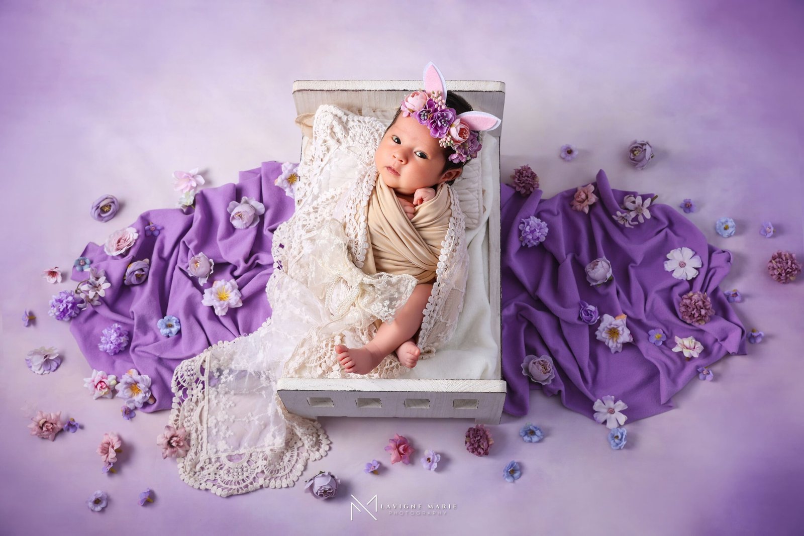 A newborn baby wrapped in beige fabric, wearing a floral headband with bunny ears, lying in a white wooden crate covered with a white lace blanket and surrounded by purple fabric and scattered artificial flowers in purple, pink, and white against a purple background